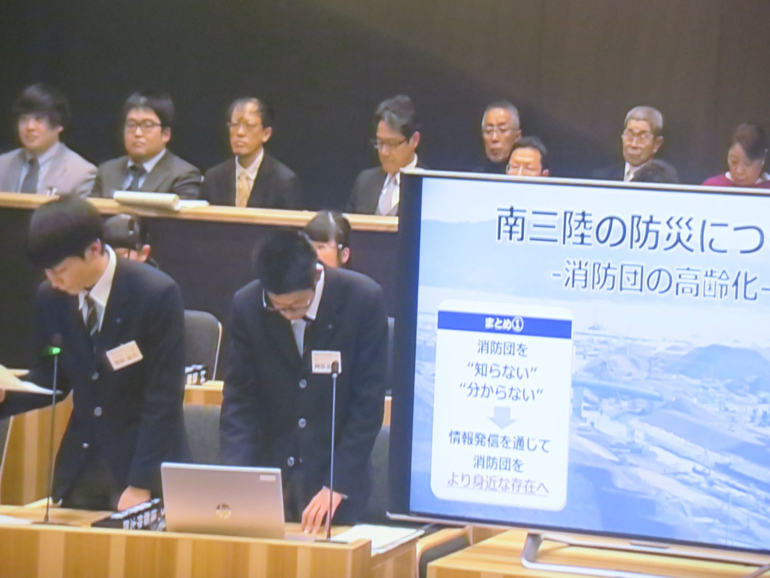 南三陸の防災に関する内容をスクリーンに映し出しながらプレゼンテーションを行っている防災グループの男子学生 2名を写した写真