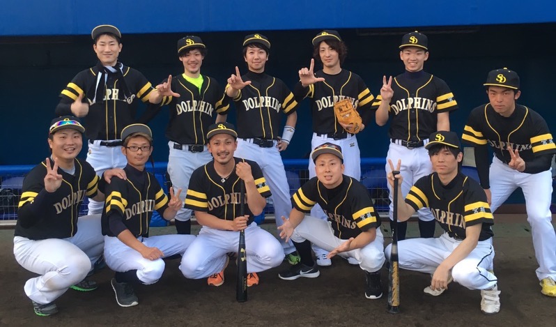 黒と黄色のユニフォームを着た草野球チーム「DOLPHINS」のメンバーたちがグラウンドのベンチ前で笑顔でポーズをとっている集合写真