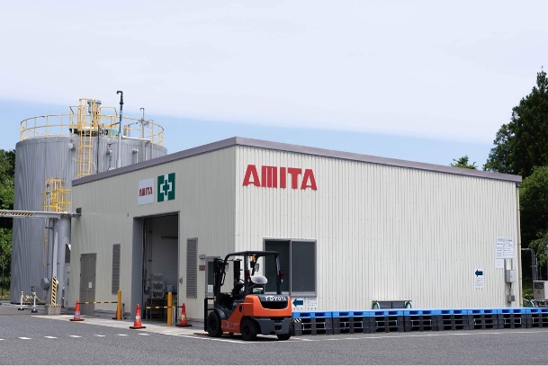 白い建物の壁に赤い文字で「AMITA」と書かれており、その手前にフォークリフト、奥には円筒形の建物が見えるバイオガス施設の外観写真