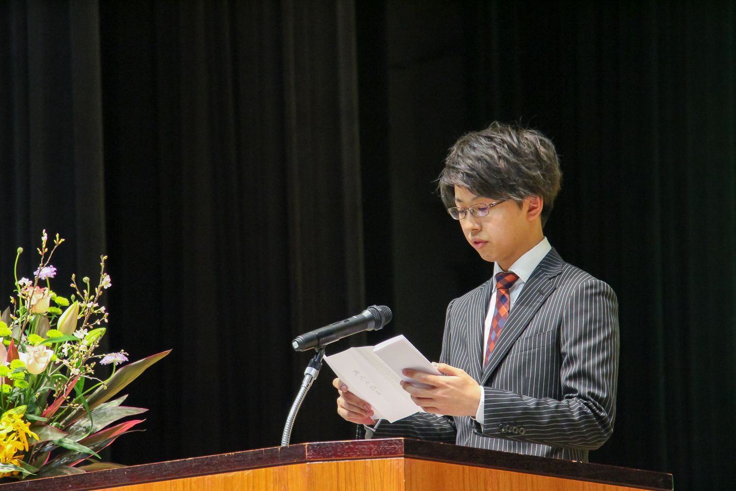 黒いスーツ姿の小野健太郎さんがマイクの前で抱負を堂々と述べている写真