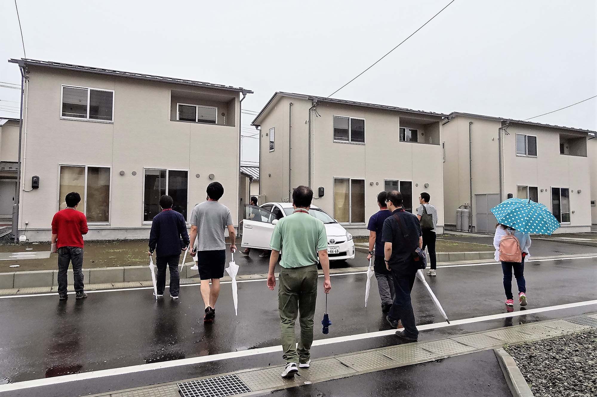 雨で濡れたアスファルトの上を、傘を持った複数の人々が住宅地に向かって歩いている様子を背後から撮影した写真