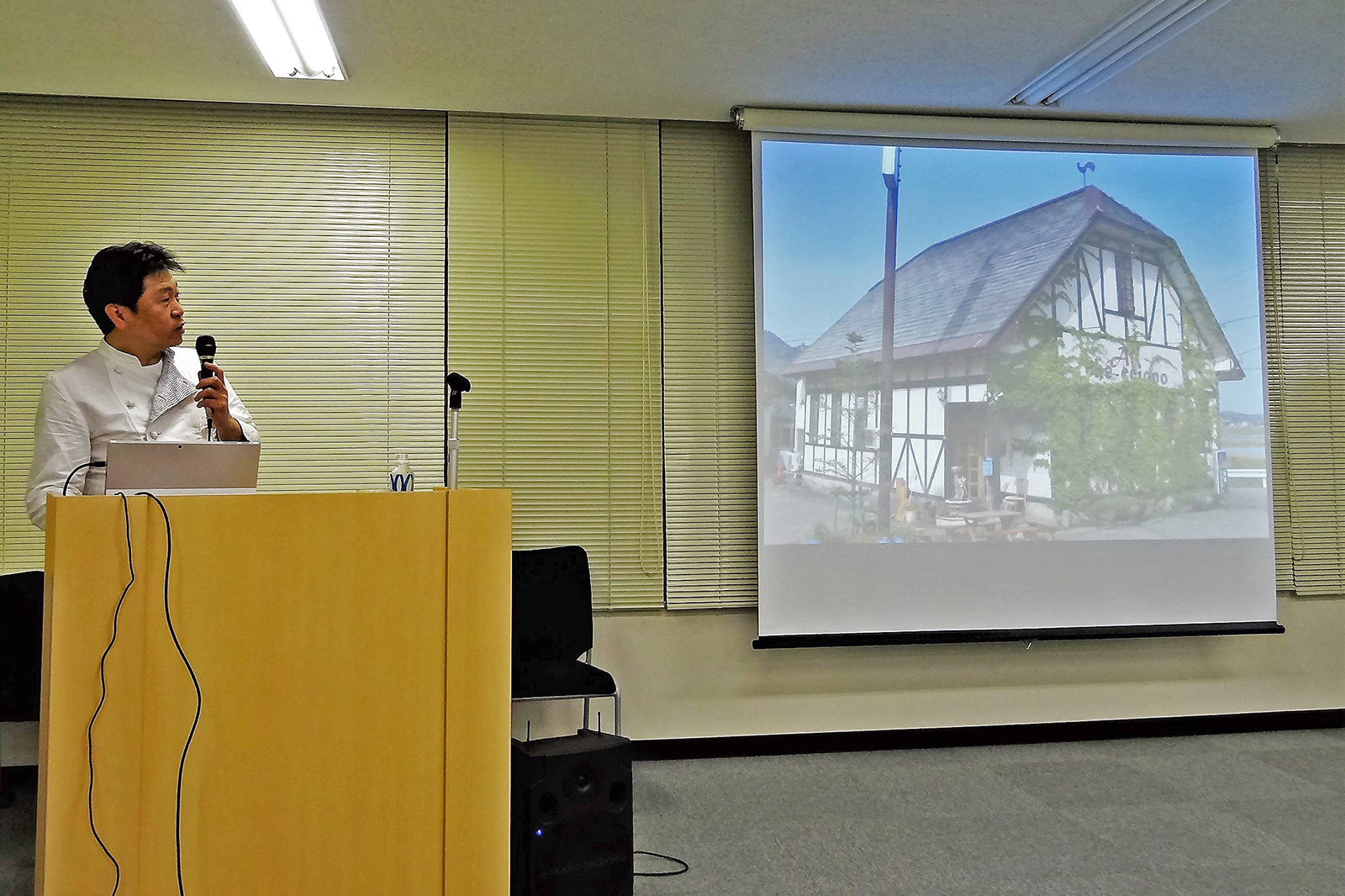 ヨーロッパ風の建物が映ったスライドの横で、演台に立ちマイクを持って話をしている奥田さんの写真