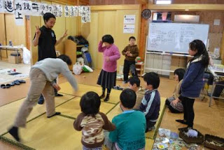 畳の上で子どもたちが円になってめんこバトルの遊びを楽しみ、笑顔のスタッフが見守っている和やかな室内の写真