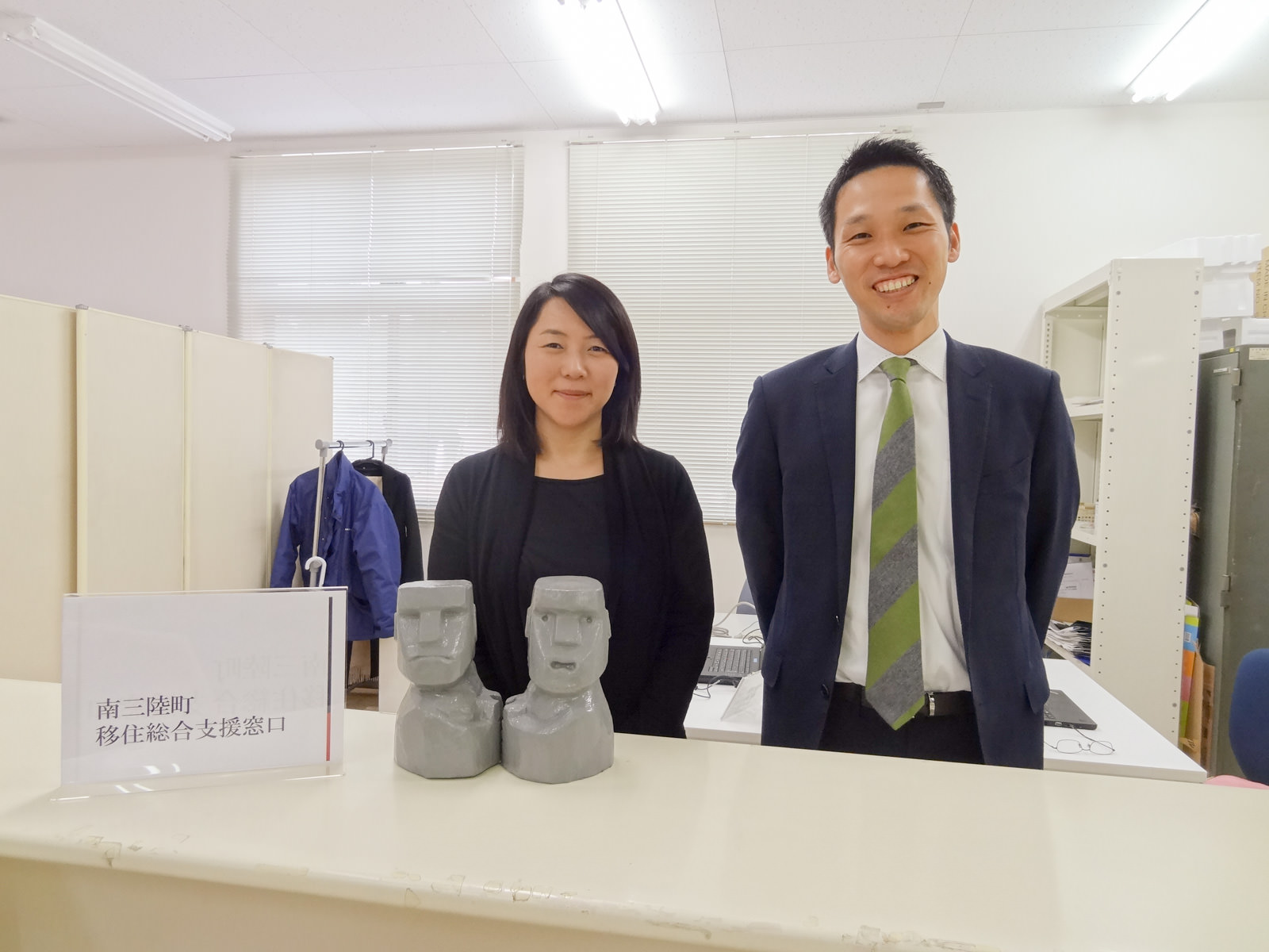 「南三陸町移住総合支援窓口」と書かれた立て札と2体のモアイ像の置物が並べて置かれた白いカウンターの後ろに、笑顔で並んで立っている片山さんと國枝さんの写真