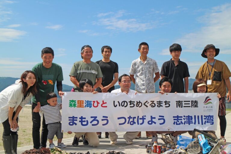 「森里海ひと いのちめぐるまち 南三陸 まもろう つなげよう 志津川湾」の横断幕を持った参加者たちの集合写真