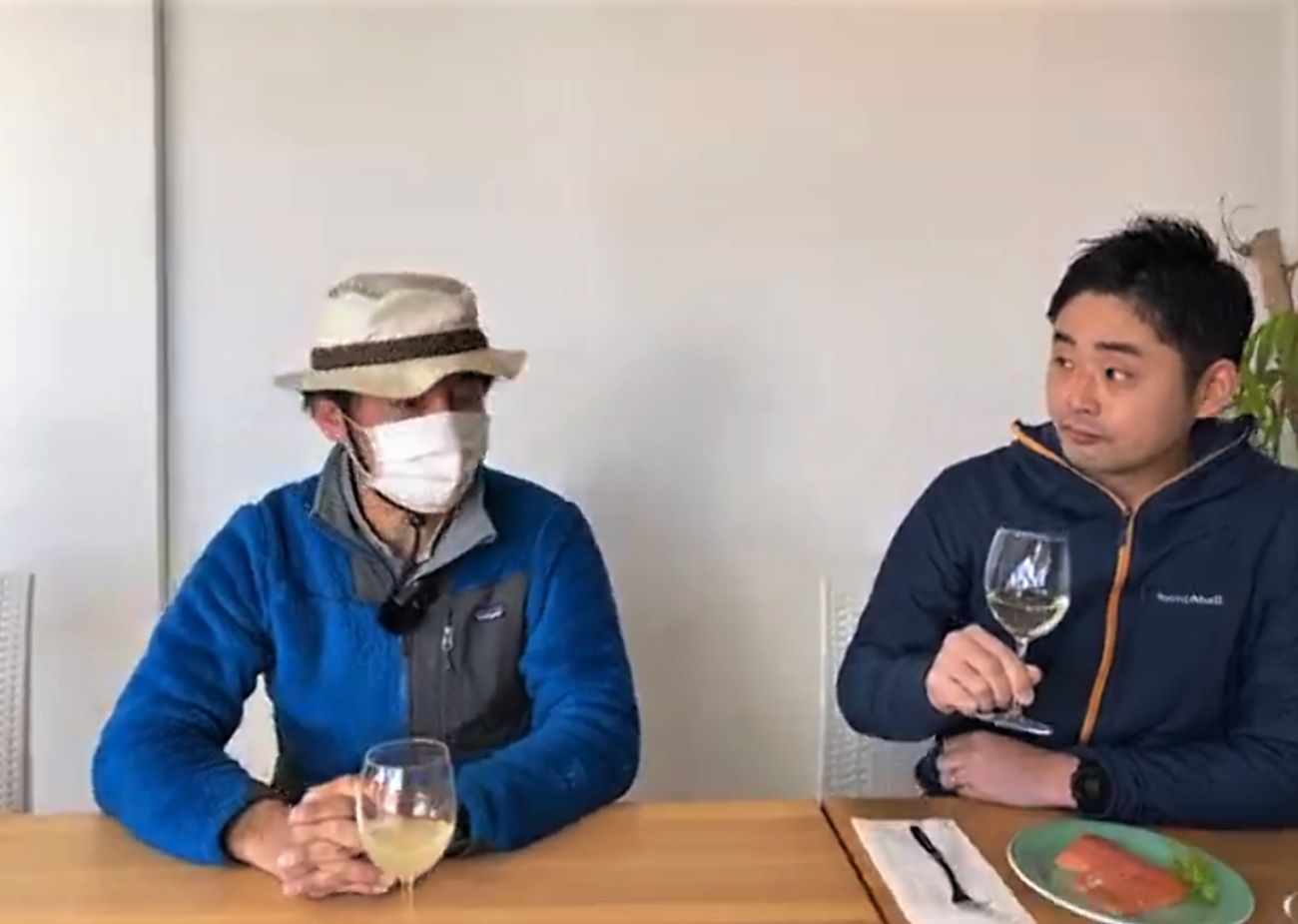 テーブルに2人の男性が座っており、左の男性は白いマスクと帽子を着けて青いフリースを着用し、右の男性は紺色のアウターを着てテーブルに置かれた銀鮭のコンフィを前にワイングラスを手にして隣の男性の話を聞いている写真