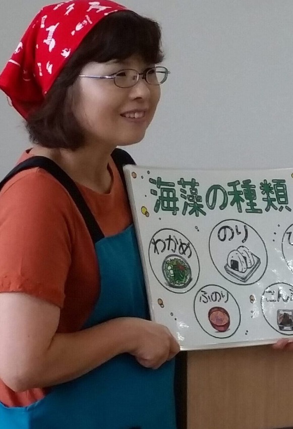 赤い三角巾と青いエプロンを着けた女性が「わかめ」「のり」「ふのり」など海藻の種類を説明するイラストボードを持ちながら、海藻について楽しそうに話している様子を写した食育講座の写真