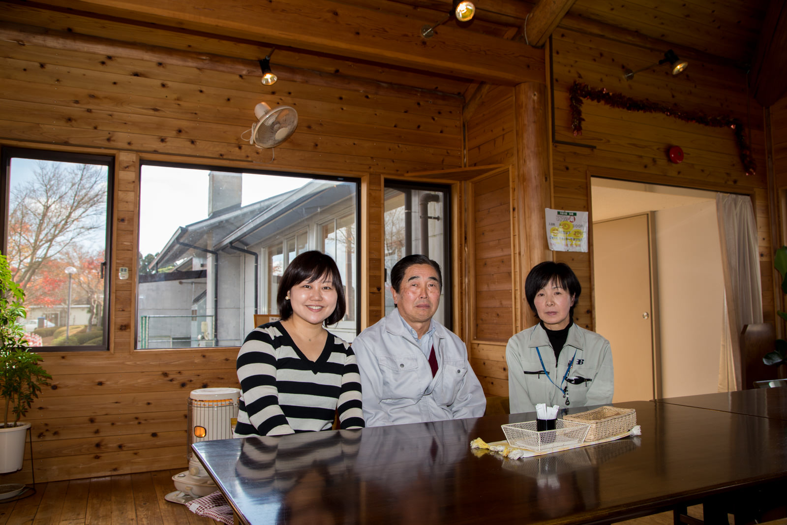 木の壁の室内に置かれた大きなテーブルの後ろに女性リポーターと男性、女性が並んで座っている写真