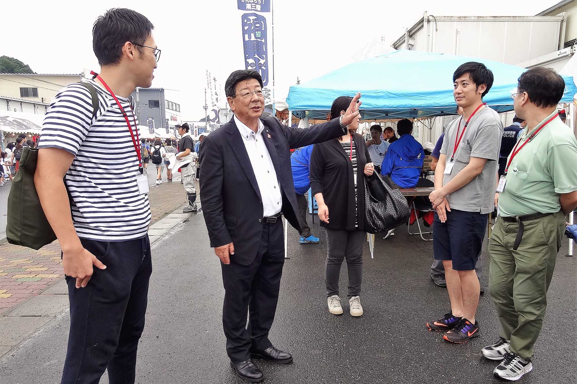 屋外イベントの場で、スーツ姿の男性が身振りを交えて話し、周囲の数人が傾聴している写真