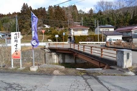 山あいの集落にある小さな橋のたもとに「そば処すがわら」の看板とのぼり旗が立っており、店の入口方向を示している写真