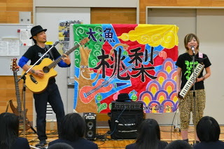 カラフルな「大魚桃梨」と書かれた幕の前で、ギターと鍵盤ハーモニカを演奏する二人組のミュージシャン、歌の上村美保子さん、ベースのJIGENさんが体育館内で歌っているミニライブの様子の写真