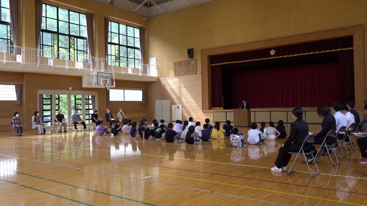 体育館のステージ前で小学生が床に座って話を聞いており、演台でスーツ姿の男性が挨拶をしていて、周囲に地域の大人たちも椅子に座って聞いている様子の写真