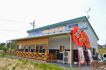 青い屋根と白い外壁の建物の前に大きな赤い開店祝いの花輪が飾られており、道路に面して木製のテラスがあるカフェの正面外観の写真