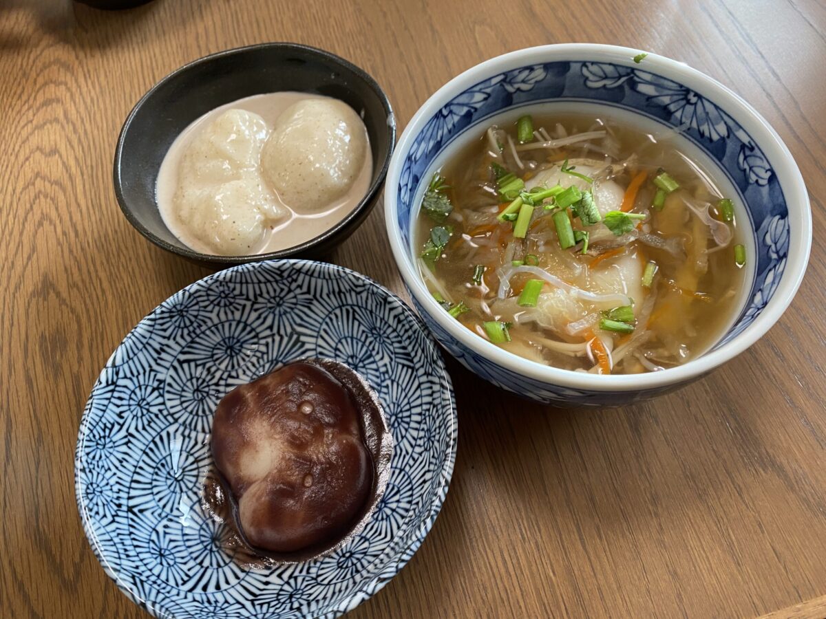 黒い器に入ったきな粉だれのかかった丸い団子が入ったくるみもちと、青い模様の器に入った小豆あんに餅が入ったあんこもち、さらに青い器に盛られた、せり、にんじん、きのこ、こんにゃくなどの具とお餅が入ったお雑煮が並んでいるお餅を使った料理の写真