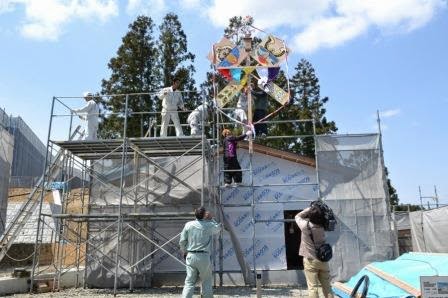 足場の上で職人たちが色とりどりの飾りを掲げ、家の棟上げを祝う準備をしている様子の写真