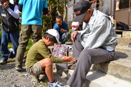 屋外の階段に座る高齢の男性が足で小さな板をしっかり押さえ、その板に地面にしゃがんだ少年が錐で丁寧に穴をあけてぶんぶんゴマを作っている様子の写真