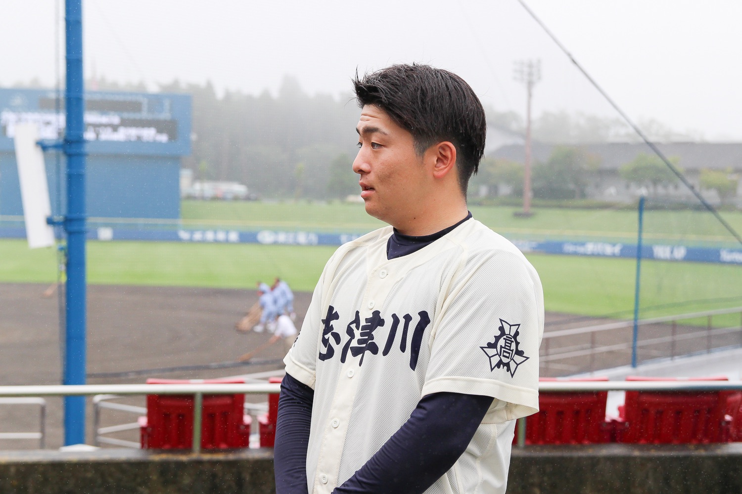 霧のかかったグラウンドを背景に、「志津川」と書かれた白いユニフォーム姿の野球選手が横向きで立っている写真