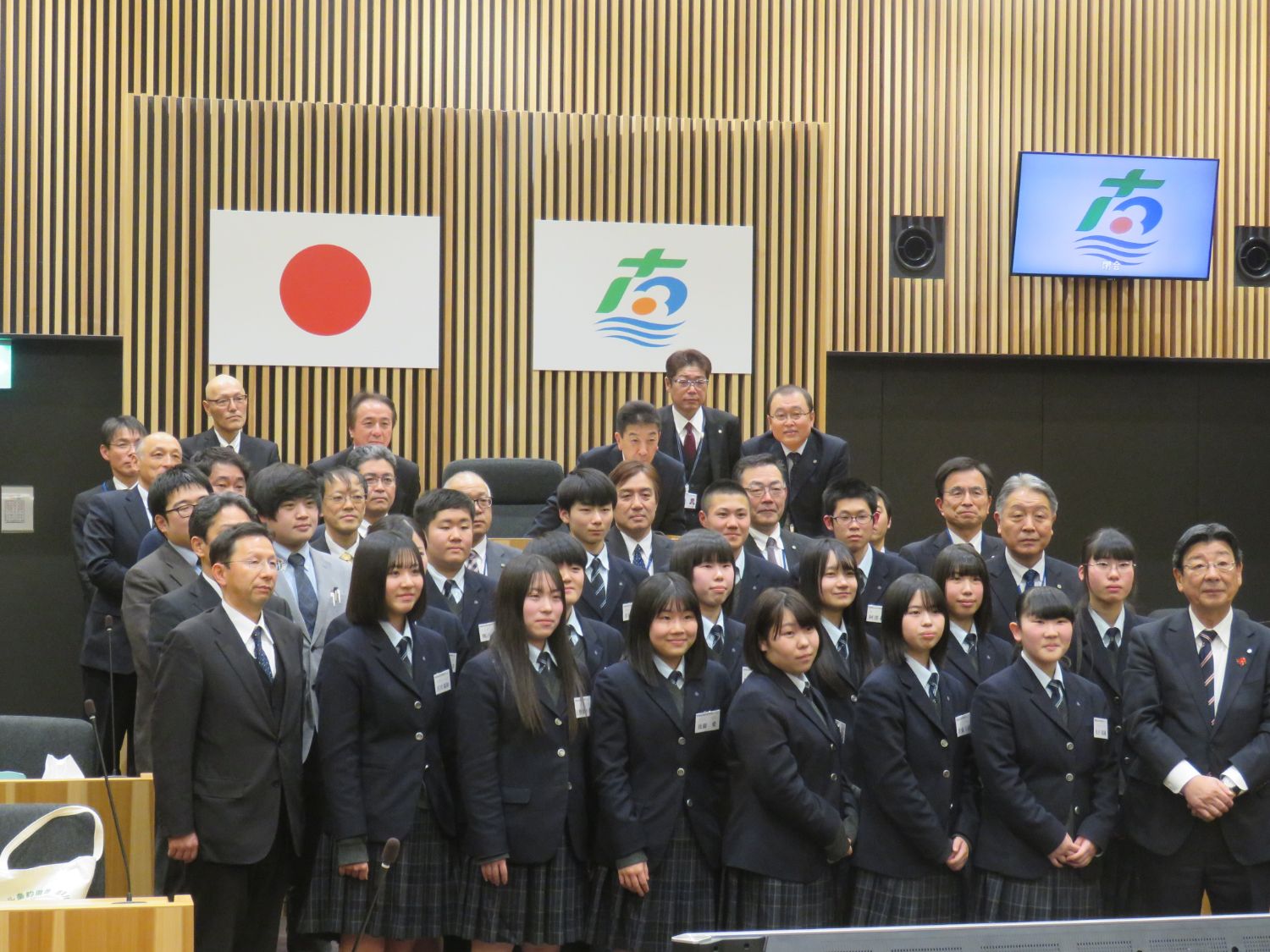 日本の国旗と町の旗を背景に、志津川高校の生徒16人と町長・副町長・各課の課長たちが整列して集合写真を撮っている写真