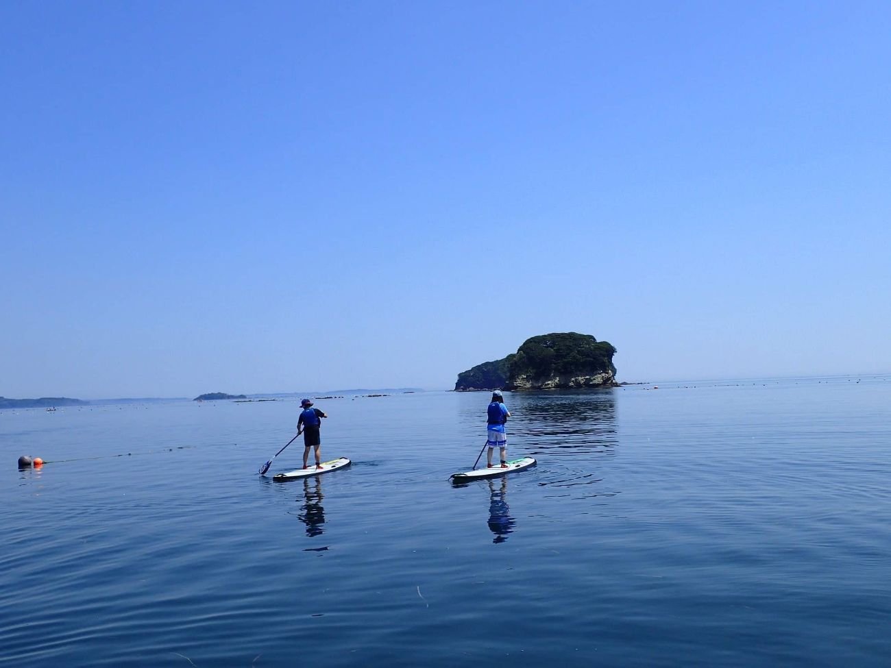 青空と水平線が広がる穏やかな海に浮かぶ小さな島を背景に、ライフジャケットを着た2人の人が立ちこぎボード(SUP)に乗ってパドルを漕いでいる様子の写真