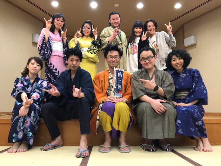 前列中央に座る男性が「南三陸」と書かれた紺色の小さな旗を持ち、前列と後列に5人ずつそれぞれ着物などの衣装を身に着けてポーズをとっている劇団員が並んでいる写真