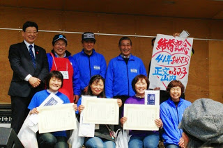 ステージの前で賞状を持った女性たちが笑顔で記念写真を撮り、後ろに青いジャンパーの男性たちが並んでいる写真