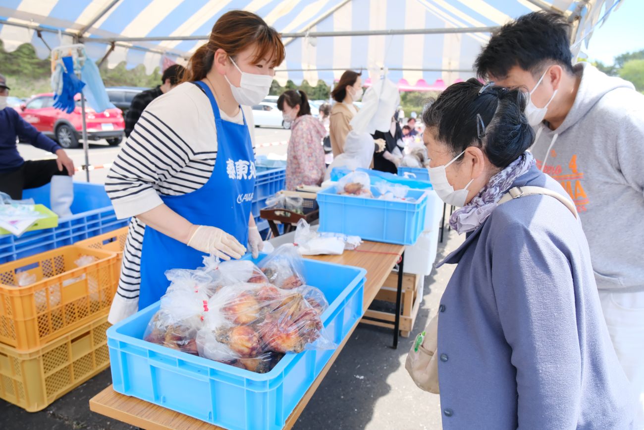 テント下の販売ブースで、手袋をした青いエプロンを着た女性が、向かい合って立つマスクをした高齢女性と男性客に、机の上に置かれた青いコンテナに入った袋詰めのホヤを販売している様子の写真