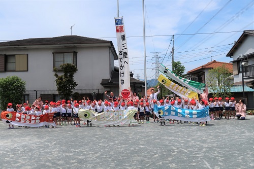 赤・緑・青の巨大な鯉のぼりを持った園児たちが園庭に集まり、先生とともに記念撮影をしている様子の写真