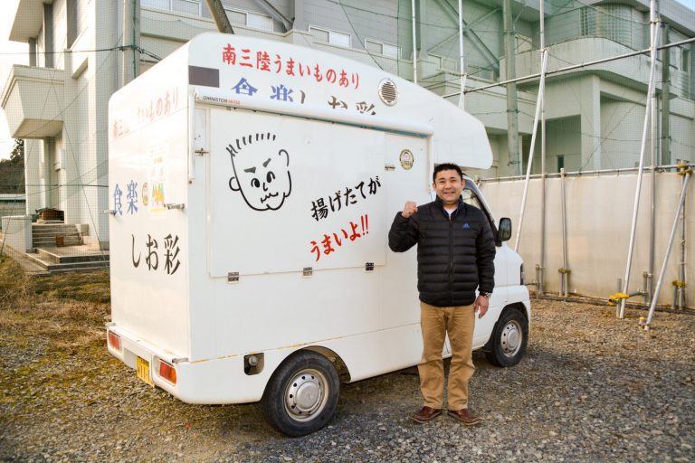 食楽しお彩と書かれ、男性の似顔絵が描かれた白い軽トラック型の移動販売車の前で、ジャンパーを着た男性が片手を握ってポーズをとっている写真
