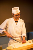 白衣と帽子を身に着けた寿司職人がまな板の上で包丁を使ってネタを切って仕込みをしている様子の写真