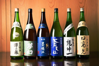 木の背景の前に「澤乃泉」「夢幻」などと書かれた地酒ラベルが貼られた6本の一升瓶が並んでいる日本酒の写真