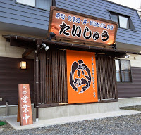 入り口正面に「たいしゅう」と書かれた看板とオレンジ色の暖簾があり、「営業中」の立て札が出ている寿司店の建物外観の写真