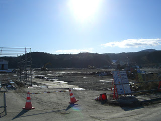 工事用のコーンと案内板の後ろに、複数の重機が見える土砂で覆われた広い土地があり、奥に山並みが広がっている写真