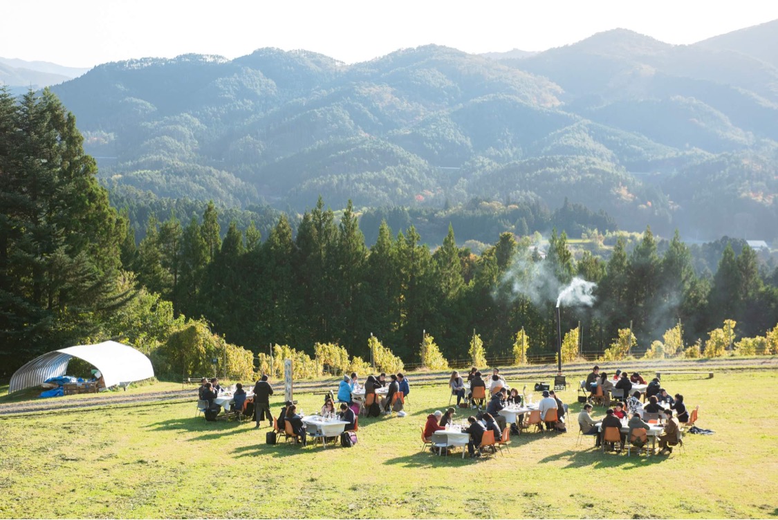 山々に囲まれた里山の高原で多くの人々が丸テーブルを囲み、青空の下で野外会食を楽しんでいる様子を写した里山の交流イベントの写真