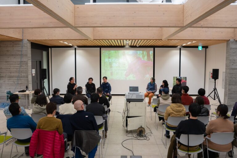 木の梁が見える室内で、参加者たちが映像が表示されたスクリーンを前に、6人の登壇者が並ぶトークショーを聴いている様子を写した写真