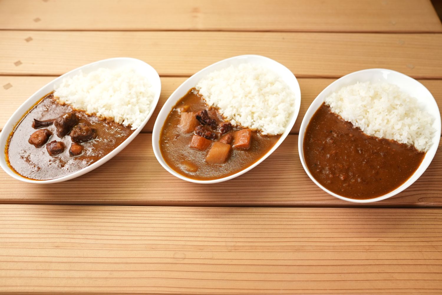 木製のテーブルの上にご飯と一緒に味の違うカレーが盛り付けられた3皿のレトルトカレーが並び、それぞれカレーの種類の違いを比較した写真