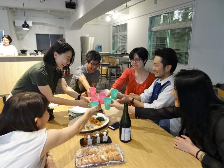 モダンなキッチンのある室内で、6人の男女が木製テーブルを囲んで料理を前にしながらカラフルなコップで乾杯している、食事会の様子の写真