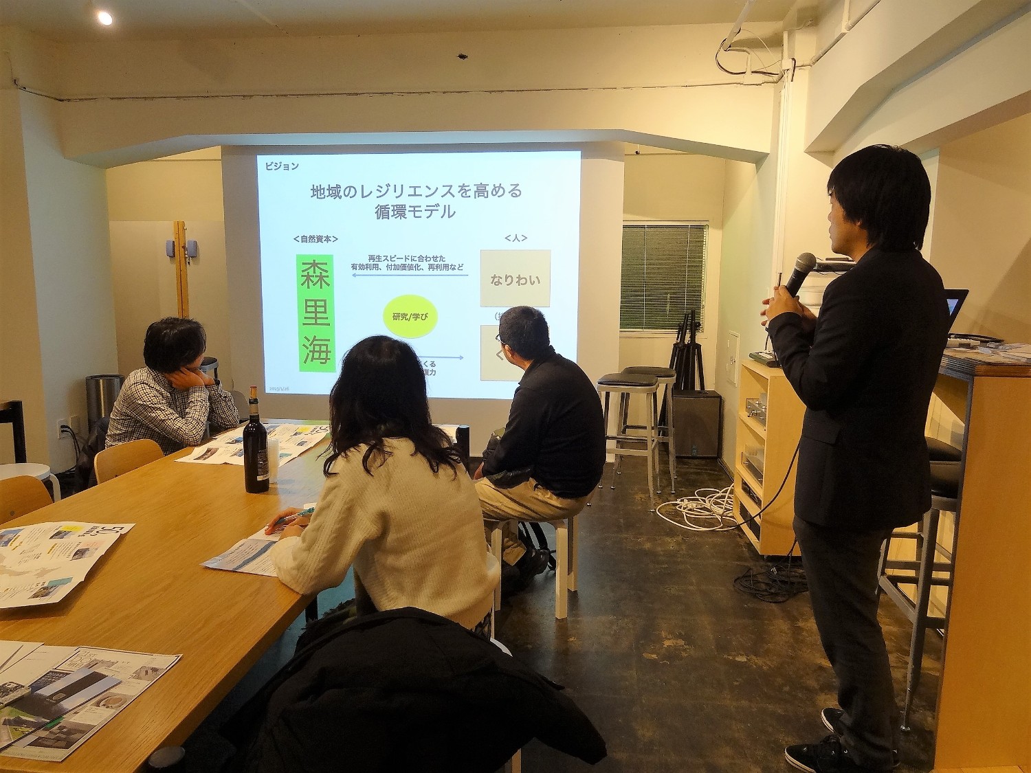 「地域のレジリエンスを高める循環モデル」と書かれたスライドが映されているスクリーンを、数人の参加者が見ながら座っており、スーツ姿の山内さんがマイクを持って話をしている写真