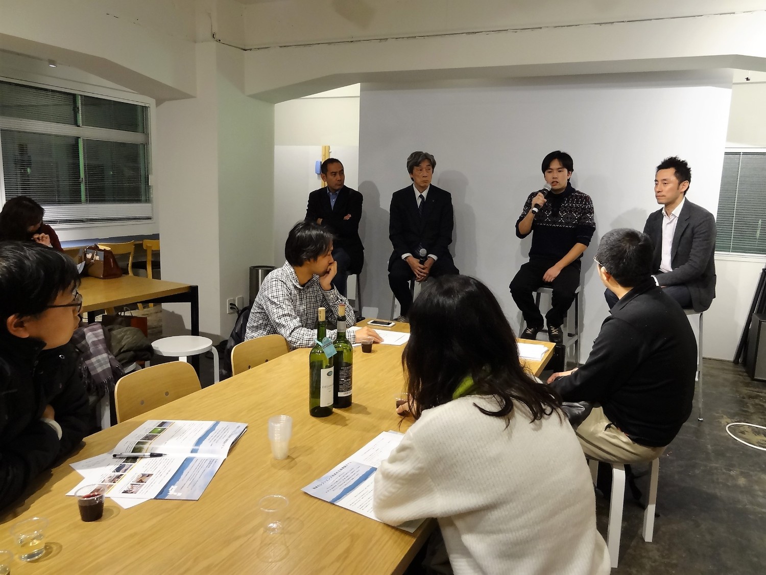 長机を囲んで数名の参加者が資料を広げて話を聞く中、白い壁の前に並んで座った4人の登壇者のうち正司さんがマイクを持って話している様子の写真