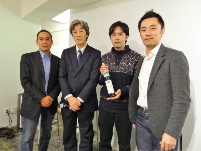 4人の男性が横一列に並び、そのうち中央の1人がワインボトルを両手で持ってカメラに向かって立っている様子の写真