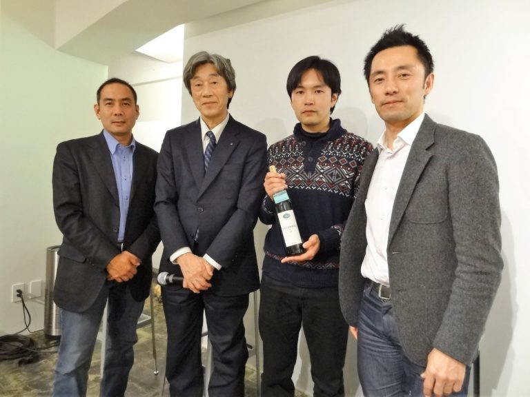 スーツ姿の男性4人が、白い壁の部屋の中でワインボトルを持ち横一列に並んでいる写真