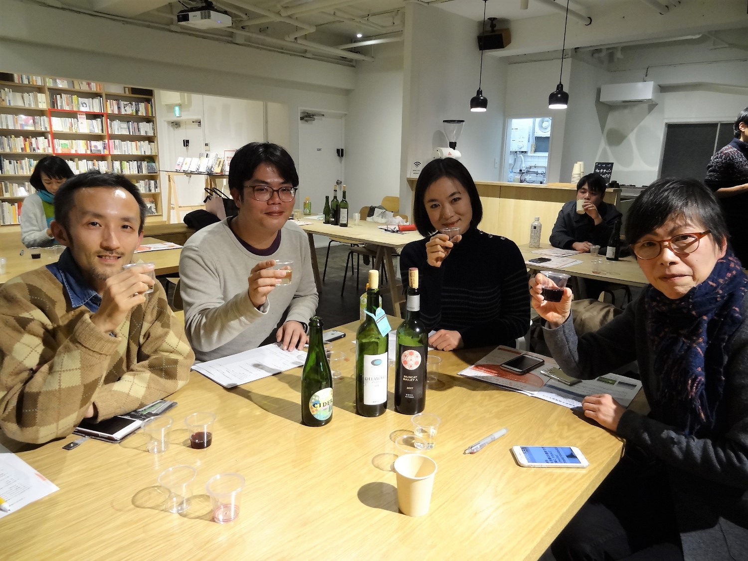 複数のワインボトルや資料が置かれているテーブルに座った4人の男女が、グラスに注がれたでワインを手に持ってカメラに笑顔を向けている写真