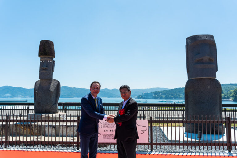 青空が広がる海の前で、2体のモアイ像の前にスーツ姿の男性と、胸に赤い胸章リボンをつけた町長が並んで立ち、互いに握手を交わしている様子を写した写真