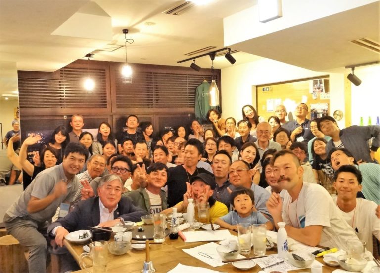 居酒屋のような室内で数十人の大人と子どもがテーブルを囲んで笑顔で集合写真を撮っている交流会の写真