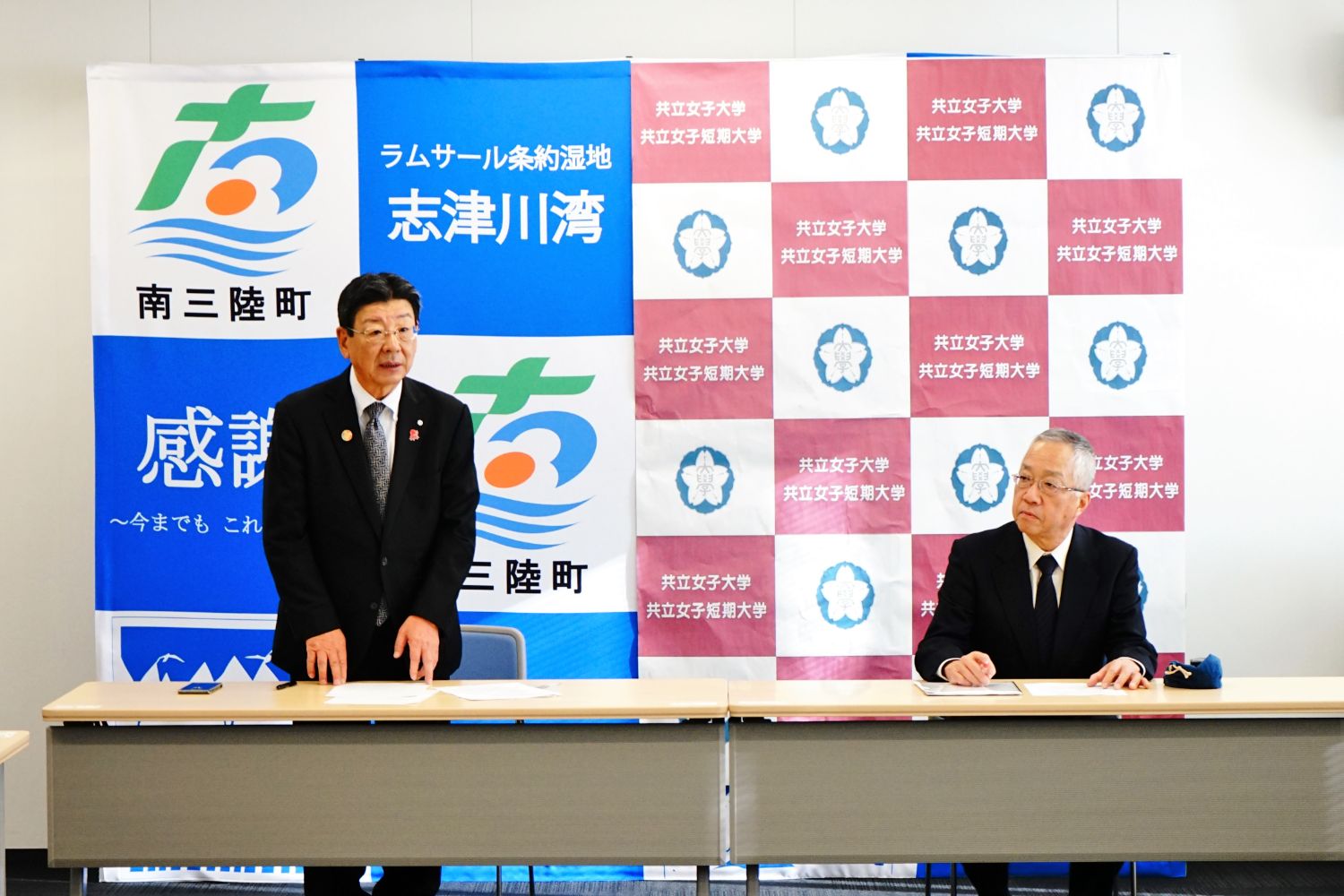 南三陸町のバックパネルの前で、スーツ姿の佐藤町長が立って挨拶をし、共立女子大学の関係者が座って話を聞いている協定締結報告会の様子の写真