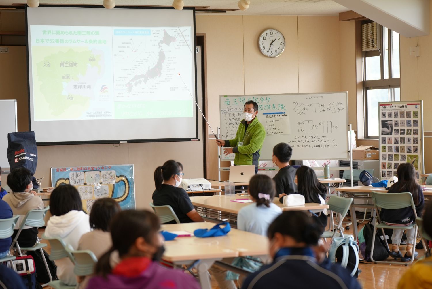 教室内でマスクをした講師が指示棒を使って大型スクリーンに写した地図を指しながら席に座った子どもたちに説明をしている様子の写真