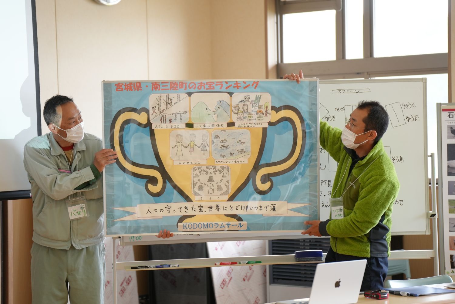 室内で二人の男性が、6枚の絵が貼られた大きなトロフィーがプリントされたパネルを掲げている写真