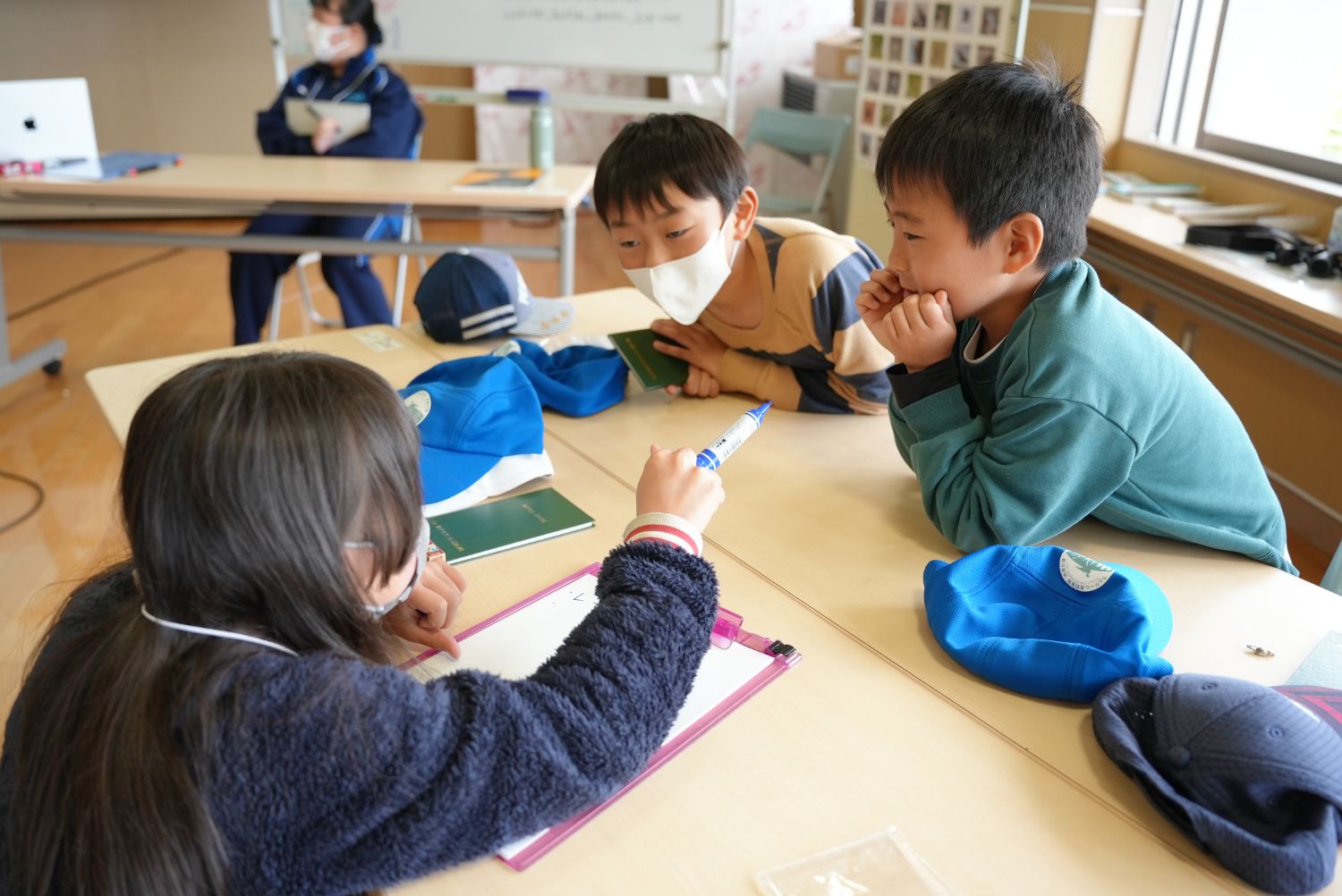 小学生3人がグループで机を囲み、真剣に話し合いをしていて、机には青い帽子やハンドブック、メモ用紙が置かれている写真