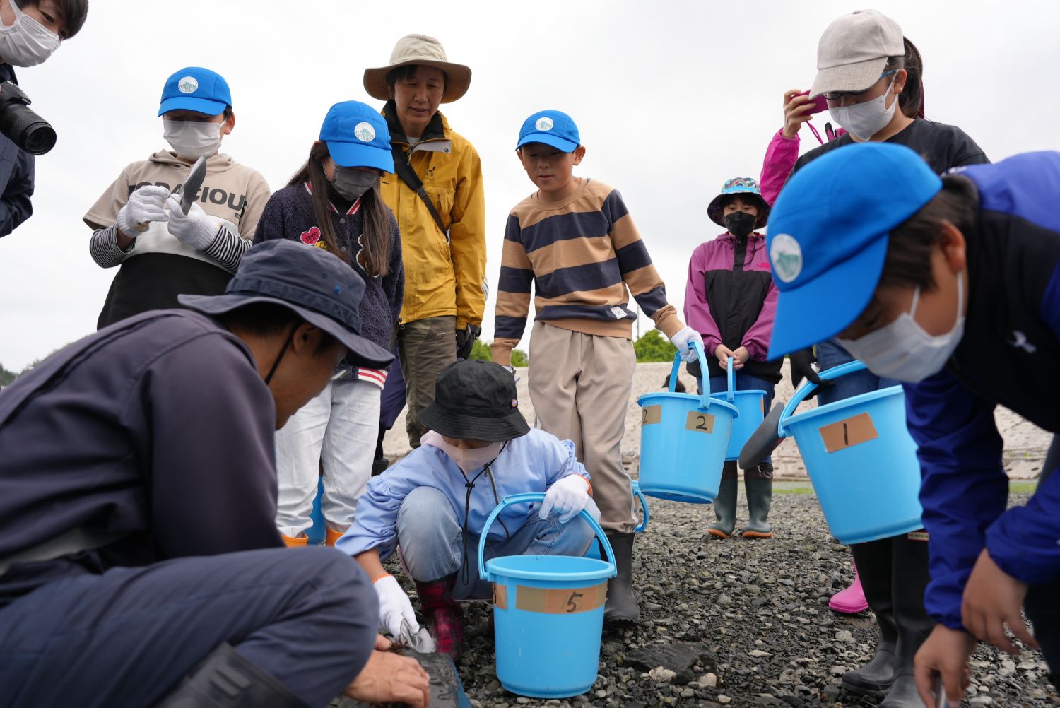 海辺で青い帽子をかぶった子どもたちが、バケツを手にスタッフの話を聞きながら地面の生き物を観察している様子を写した写真