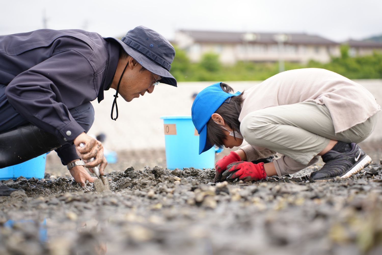 青いバケツを横に置いた児童と大人が、しゃがんで地面に顔を近づけ、熱心に調査している様子の写真