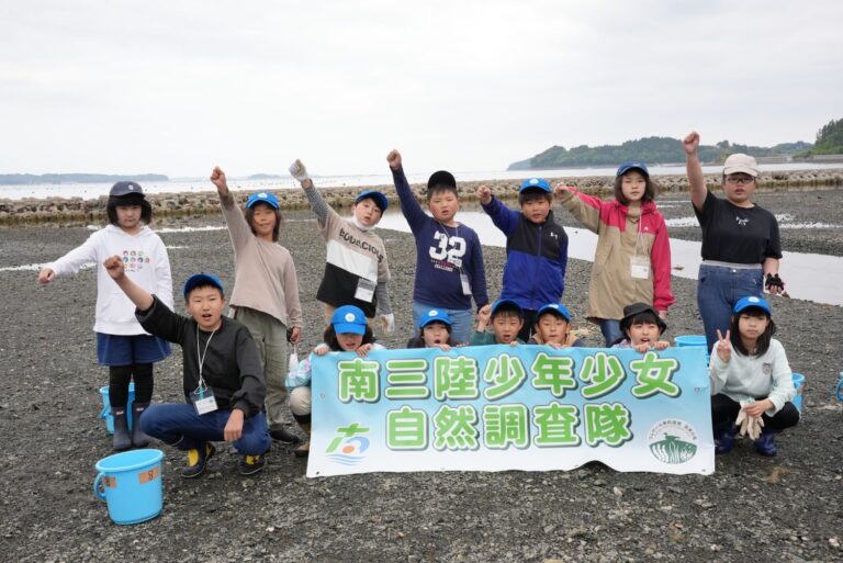 海岸で「南三陸少年少女自然調査隊」と書かれた横断幕を掲げながら、メンバー達が並び右手を上げている集合写真